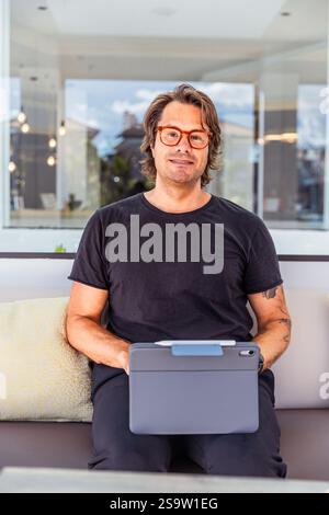 Vertical portrait of a man in a black shirt is sitting on a couch holding a tablet. Stock Photo