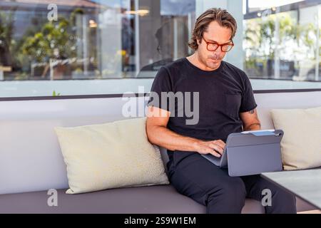 A concentrated man is sitting on a couch working on his tablet, photo with copy space. Stock Photo