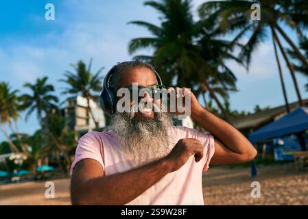 Senior man with gray beard wearing headphones dances on tropical beach. Enjoys music under palm trees on summer day relishing vibrant tunes. Perfect Stock Photo