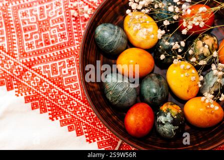 Multicolored Easter eggs in clay bowl on embroidered red towel. Ukrainian various embroidery techniques. Dyeing Easter eggs with hibiscus. Stock Photo