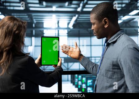 System administrators in data center use green screen tablet to calibrate energy usage. Teamworking server farm colleagues using mockup device to inspect UPS configurations, doing maintenance on gear Stock Photo