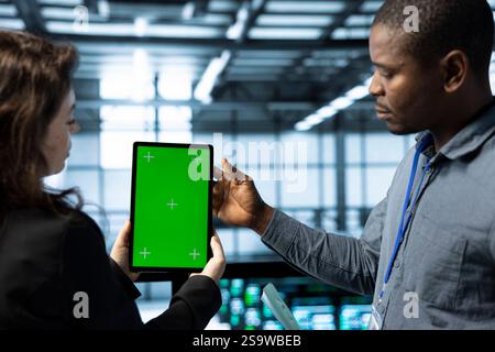 Engineers in data center using mockup tablet, observing analytics, reviewing supercomputer gear health. Server hub IT colleagues chatting, using chroma key device, adjusting system parameters Stock Photo