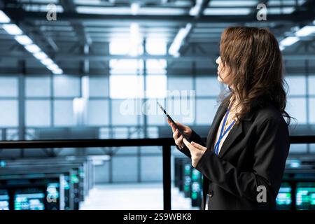 Programmer in data center facility uses tablet, configuring rigs network virtualization. IT expert in server farm updating storage arrays with device, verifying settings to maintain stable performance Stock Photo