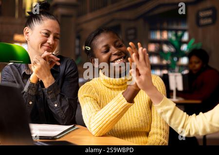 Diverse teen scholars doing high five gesture together at the end of tutoring session with mentor, teacher helping the girls with accurate learning materials in the library. Mentoring pupils. Stock Photo
