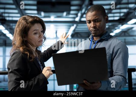 Computer scientists in data center automating routine maintenance tasks with laptop, executing diagnostic scripts. Server farm IT experts doing brainstorming, using device, adjusting power settings Stock Photo