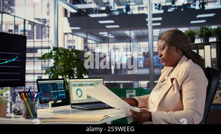Black influential businesswoman evaluates company performance, using data reports and analytics to make informed decisions that optimize efficiency and productivity. Work ethic. Camera B. Stock Photo