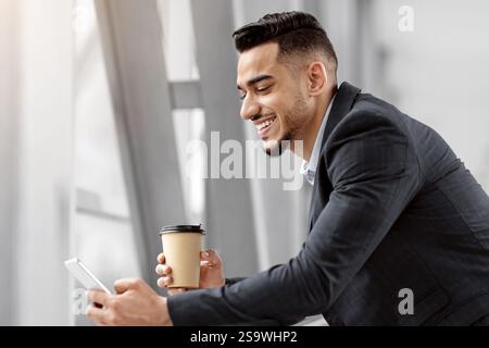 Happy Middle-Eastern Businessman Using Smartphone And Having Coffee While Waiting In Airport Stock Photo