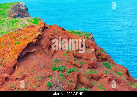 Vibrant coastal landscape featuring rugged red cliffs with patches of green vegetation, set against the backdrop of a bright blue ocean. The contrast Stock Photo