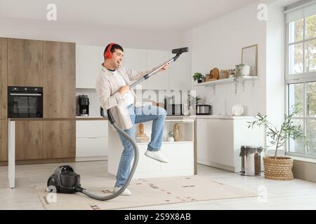 Young man having fun and listening to music while vacuuming carpet in kitchen Stock Photo