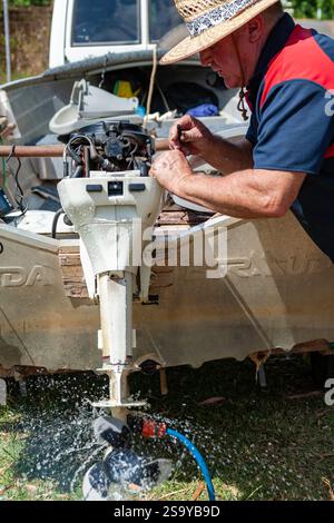 man performs maintenance outboard motor small boat scene demonstrates hands on work repair outdoor activity water spraying off propeller straw hat Stock Photo