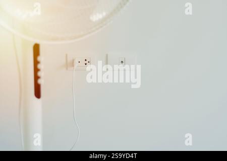 Cropped shot of white modern electric fan in light room with power plug on wall background. Selective focus on plug. Stock Photo