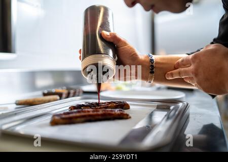 A chef carefully drizzling soy sauce over grilled eel fillets on a tray in a professional kitchen, emphasizing culinary art and food preparation. Stock Photo
