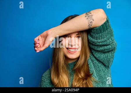 Studio portrait of a cheerful young woman with long hair and tattoo, wearing a green sweater, hiding her eyes with her arm, against a vibrant blue bac Stock Photo