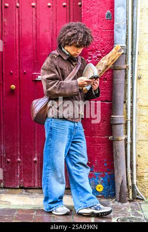 Young, casually dressed man with frizzy hair, standing in doorway holding bread and checking phone - Tours, Indre-et-Loire (37), France. Stock Photo