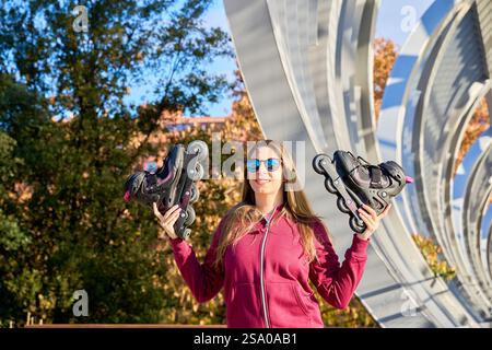 Smiling woman holding roller skates in a vibrant urban park, enjoying a sunny day filled with fun and freedom while embracing an active lifestyle Stock Photo