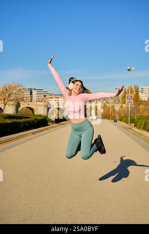 Energetic young woman wearing headphones enjoying a sunny day while jumping with inline skates in an urban park, expressing freedom and happiness Stock Photo