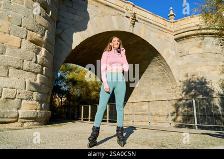 Sporty woman wearing leggings and top, holding sunglasses, posing with inline skates under a stone bridge arch on a sunny day Stock Photo