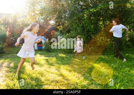 A joyful family moment as a mother and her two daughters run together in a lush green backyard on a sunny summer day, laughing and playing amidst natu Stock Photo