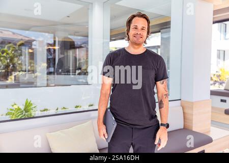 A businessman casually dressed is holding a tablet and smiling. He is standing in front of a window with a couch in the background Stock Photo