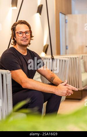 A man in a black shirt and glasses sits on a couch, vertical photo. He is smiling and he is relaxed Stock Photo
