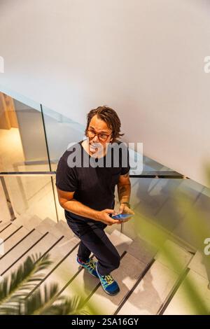 A man wearing glasses and a black shirt is walking up a set of stairs while holding a cell phone Stock Photo