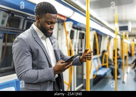 Smiling businessman holding a pole while using a smartphone, standing inside a subway train during his daily commute in the city Stock Photo