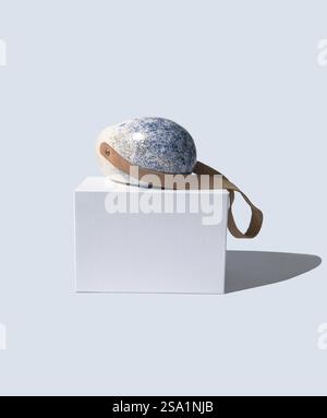 Textured rock with leather strap on white cube against light background Stock Photo