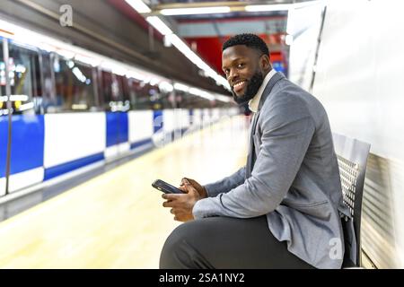 Young black businessman sitting on bench in subway station using smartphone while waiting for train Stock Photo