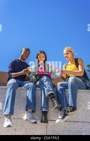 Vertical low angle view photo with copy space against blue sky of multi-ethnic students sitting on a wall outside the campus using mobile phones Stock Photo