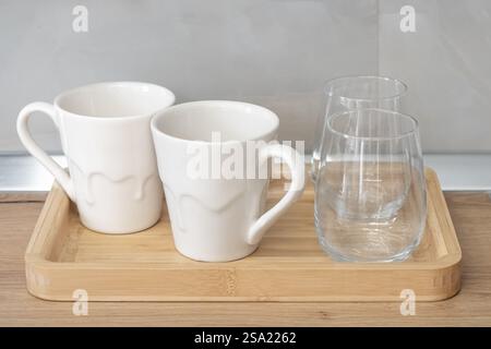 White cups for coffee or tea and drinking glasses. Two empty water glasses and two tea cups or mugs on a wooden tray in the kitchen, close up. Table w Stock Photo