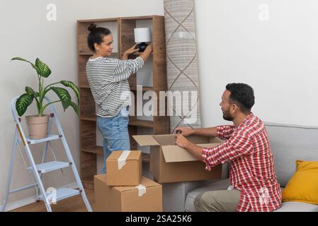 cheerful couple unpacks and arranges a lamp empty shelf in new home, relocation Stock Photo