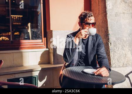 Young stylish man relaxes in cafe outdoor drinking coffee on terrace. Businessman enjoys beverage during coffee break on street Stock Photo