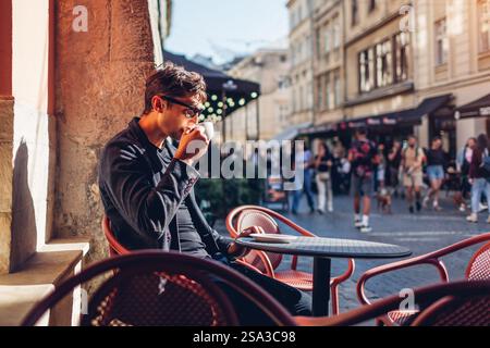 Portrait of stylish man relaxing in outdoor cafe drinking coffee. Businessman enjoying architecture of European city sitting at table on street alone Stock Photo