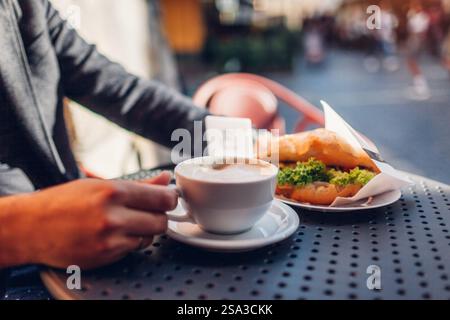 Close up of cup of coffee with croissant in male hands. Man drinking hot cappuccino beverage in outdoor cafe at sunset sitting at table on terrace. Stock Photo