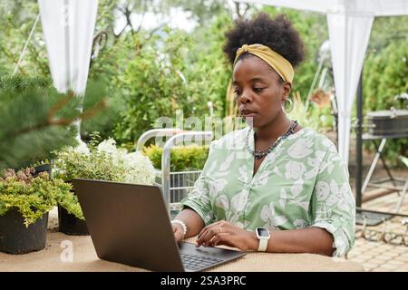 Chest up shot of focused African American female landscape designer typing on laptop, while working on garden project online sitting at table with lively summer greenery outdoors Stock Photo