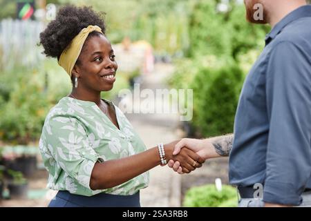 Medium shot of cheerful African American female landscape designer shaking hands with male partner making successful business deal while meeting outside in plant nursery, copy space Stock Photo