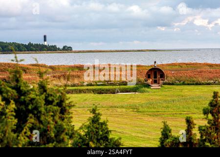 A serene coastal view showcasing a unique barrel-shaped sauna amidst vibrant fall colors, with a lighthouse in the backdrop and calm waters providing Stock Photo