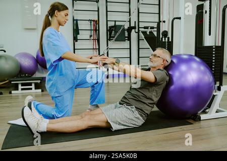 Side view of young female rehab specialist doing exercise with elderly man suffering from injure, he sitting on mat and leaning on ball Stock Photo