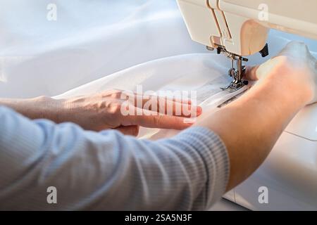 Person using a sewing machine to stitch fabric. Close-up of hands working on a sewing project with white material. Stock Photo