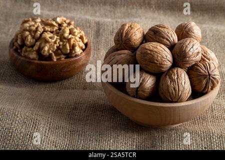 A wooden bowl filled with walnut kernels, placed alongside whole walnuts in their shells on burlap fabric Stock Photo