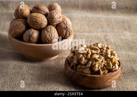 A wooden bowl filled with walnut kernels, placed alongside whole walnuts in their shells on burlap fabric Stock Photo