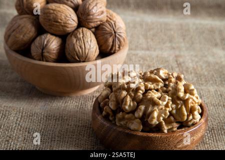 A wooden bowl filled with walnut kernels, placed alongside whole walnuts in their shells on burlap fabric Stock Photo