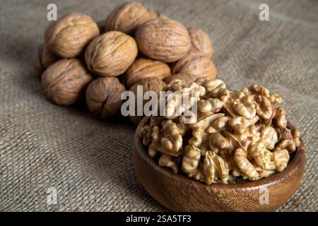 A wooden bowl filled with walnut kernels, placed alongside whole walnuts in their shells on burlap fabric Stock Photo