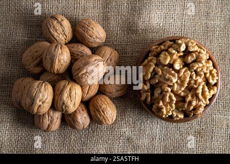 A wooden bowl filled with walnut kernels, placed alongside whole walnuts in their shells on burlap fabric Stock Photo