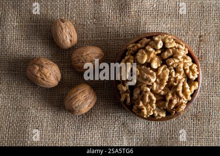 A wooden bowl filled with walnut kernels, placed alongside whole walnuts in their shells on burlap fabric Stock Photo
