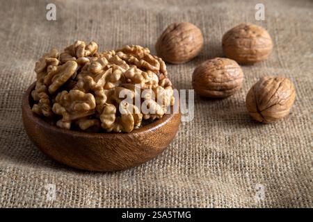 A wooden bowl filled with walnut kernels, placed alongside whole walnuts in their shells on burlap fabric Stock Photo