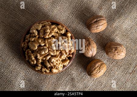 A wooden bowl filled with walnut kernels, placed alongside whole walnuts in their shells on burlap fabric Stock Photo