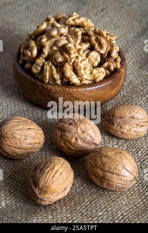 A wooden bowl filled with walnut kernels, placed alongside whole walnuts in their shells on burlap fabric Stock Photo