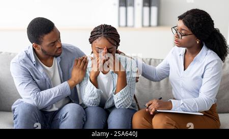 Young black woman crying at therapist's office, affectionate husband supporting her with professional psychologist Stock Photo