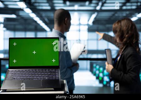Green screen laptop in data center with programmers in blurry background employing machine learning models. Close up of chroma key notebook used by teamworking server room IT experts Stock Photo